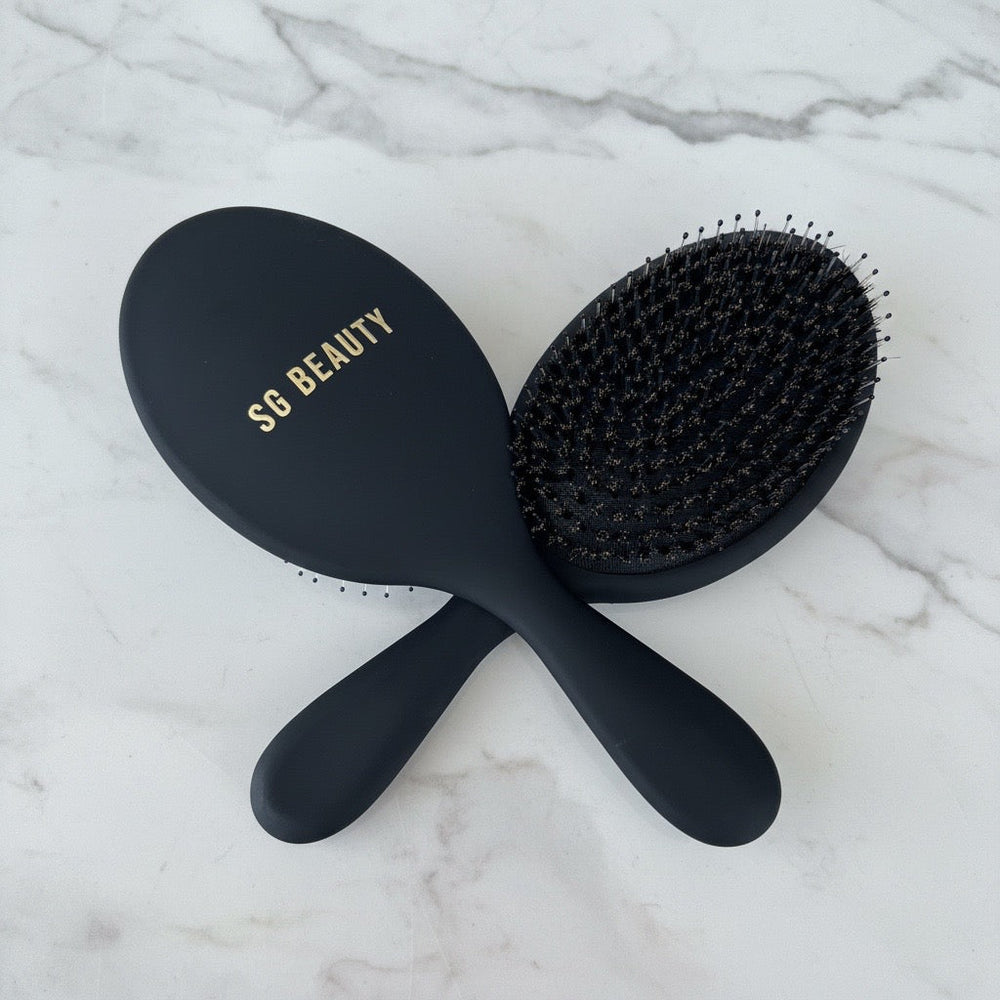Two black hairbrushes crossed on a white marble surface, with one brush showing the oval head and gold 'SG BEAUTY' branding and the other showing the bristle side.