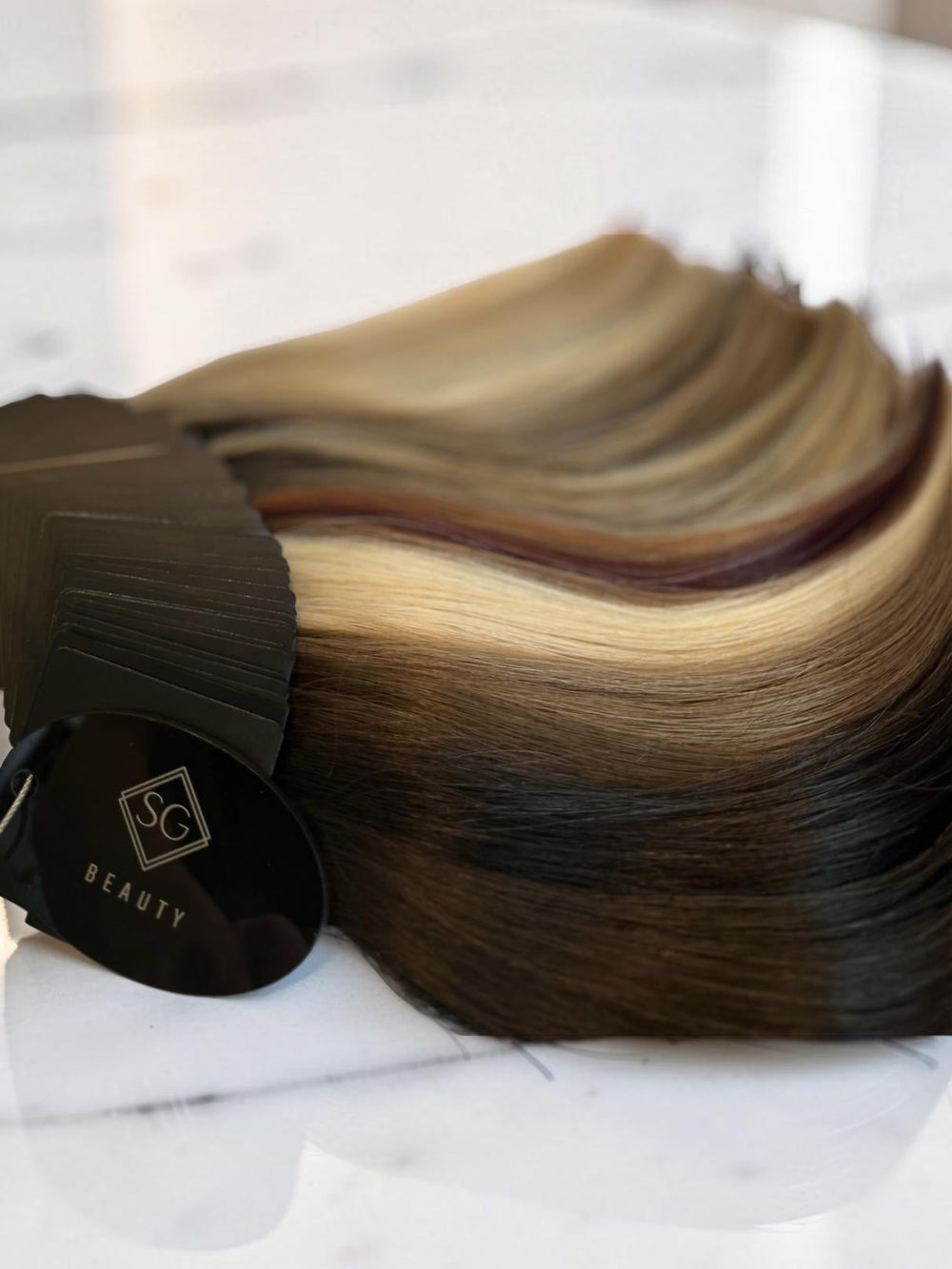 A close-up of a hair color sample fan with dark brown, black, and blonde streaks, featuring a black tag with the SC BEAUTY logo.